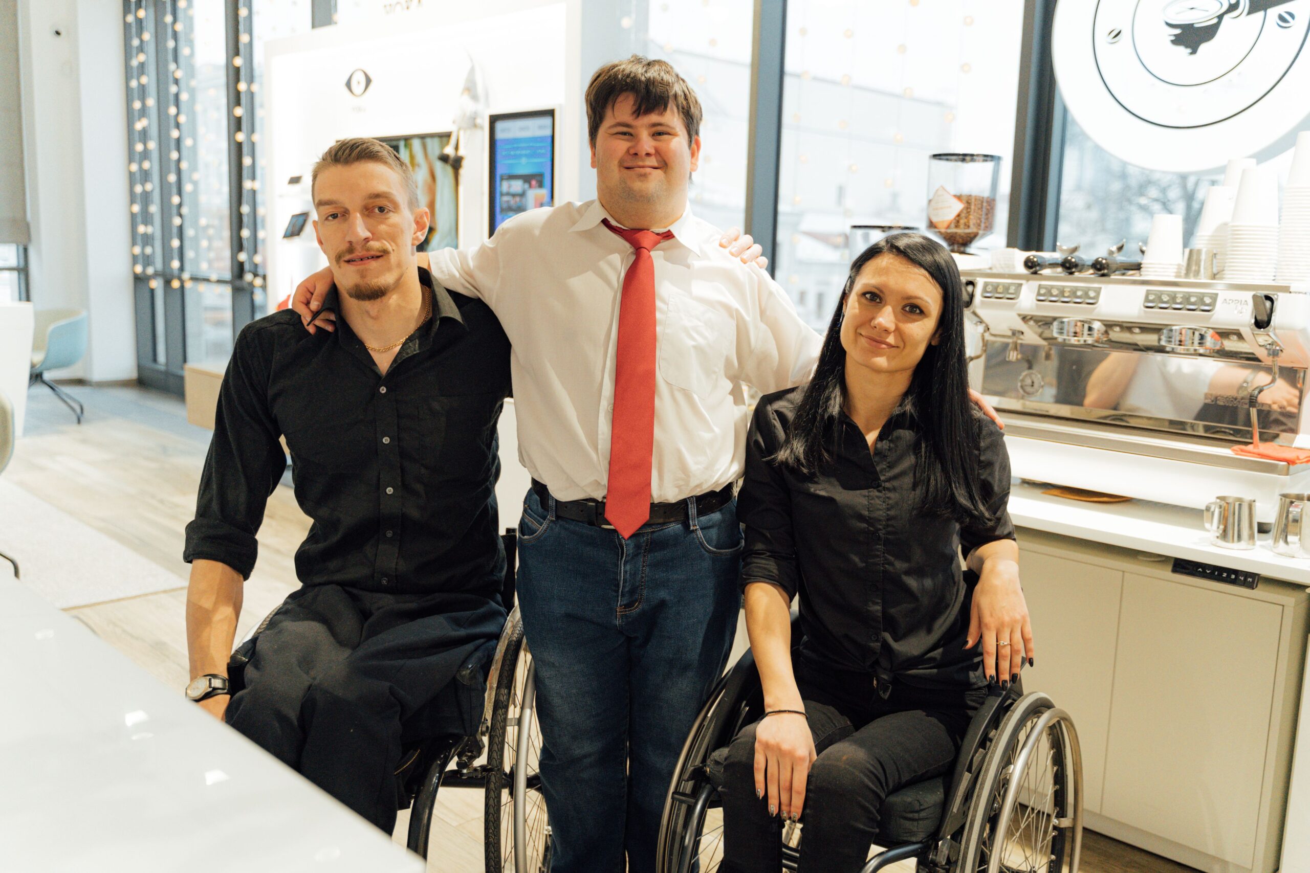 Three baristas, including two in wheelchairs, smiling at a coffee shop. Promoting inclusivity.